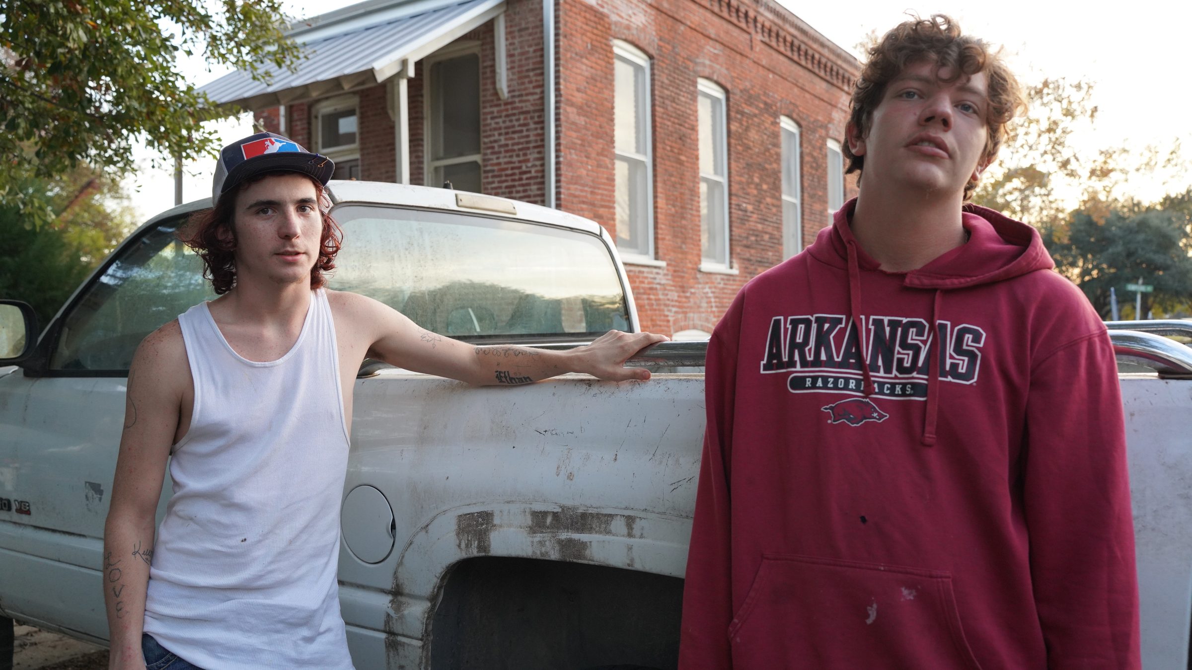 Two young men standing beside a weathered white pickup in front of a brick building.