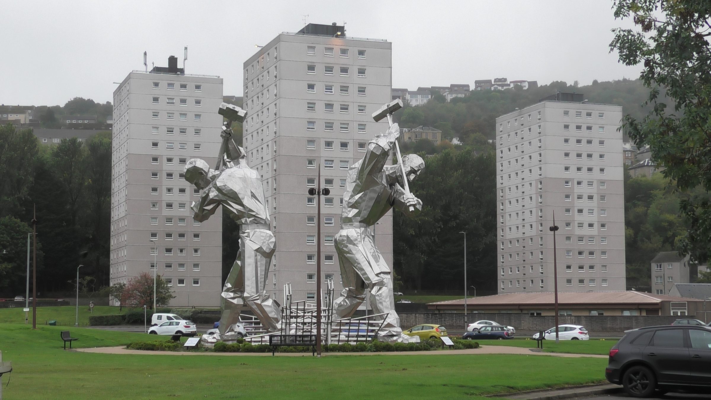 Two silver hammer-wielding statues in front of high-rises on a grey Scottish hillside.