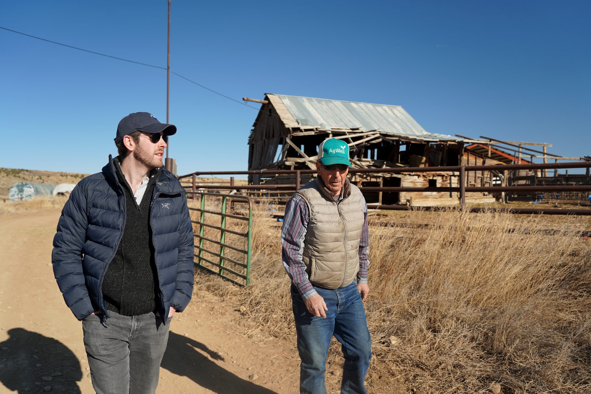 A Roca correspondent walking a ranch road with a local source, a collapsing barn behind them under a clear blue sky.
